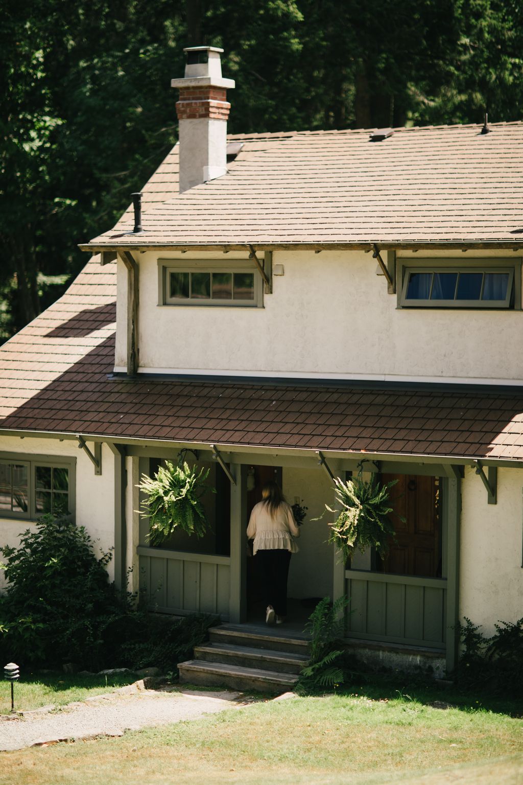 Woman entering a sage-green cottage entrance framed by hanging ferns, with shingled roof and brick chimney rising above