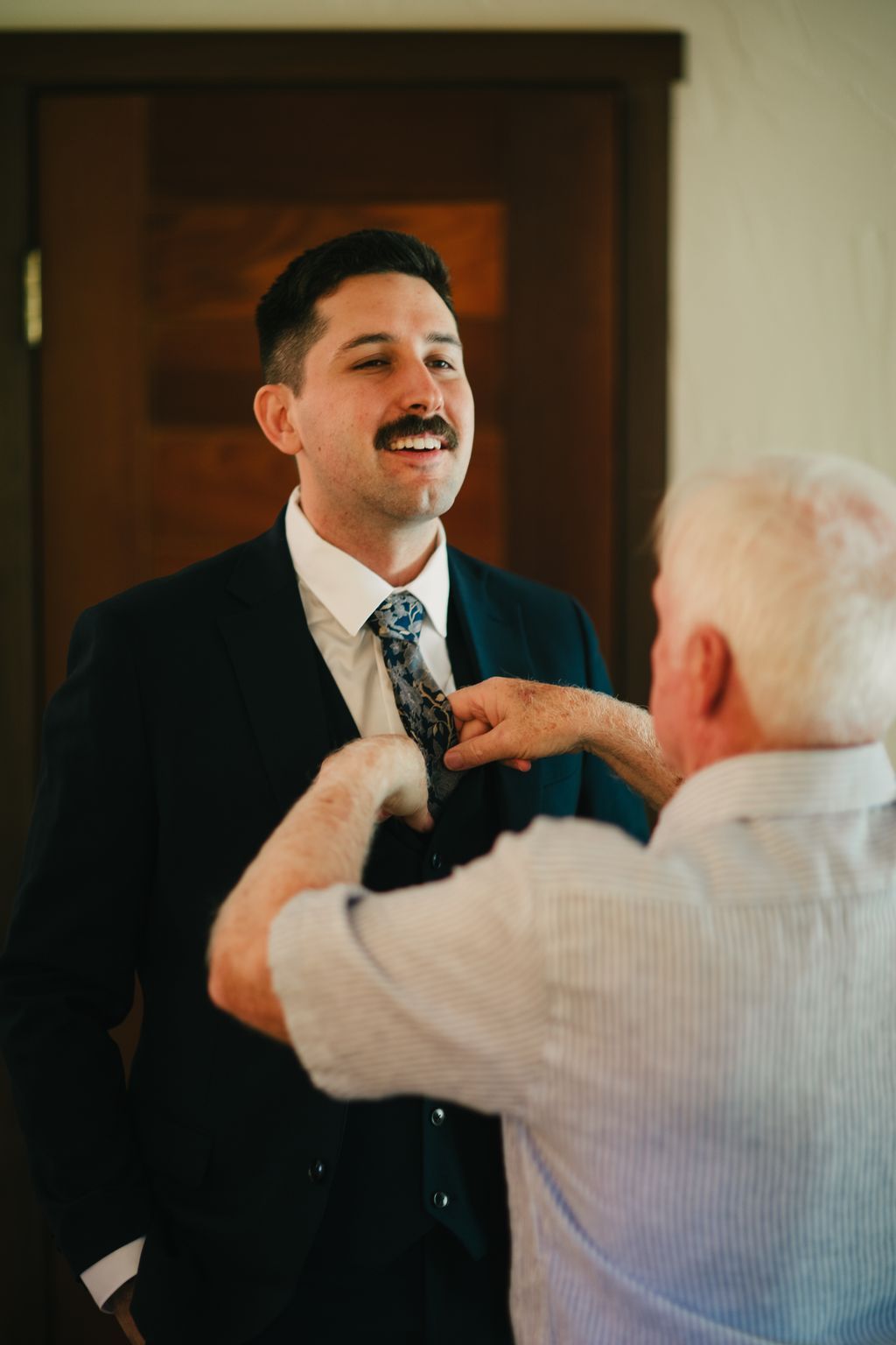 Groom in navy blazer and patterned tie smiles while someone adjusts his tie, warm interior light catching the moment before the ceremony