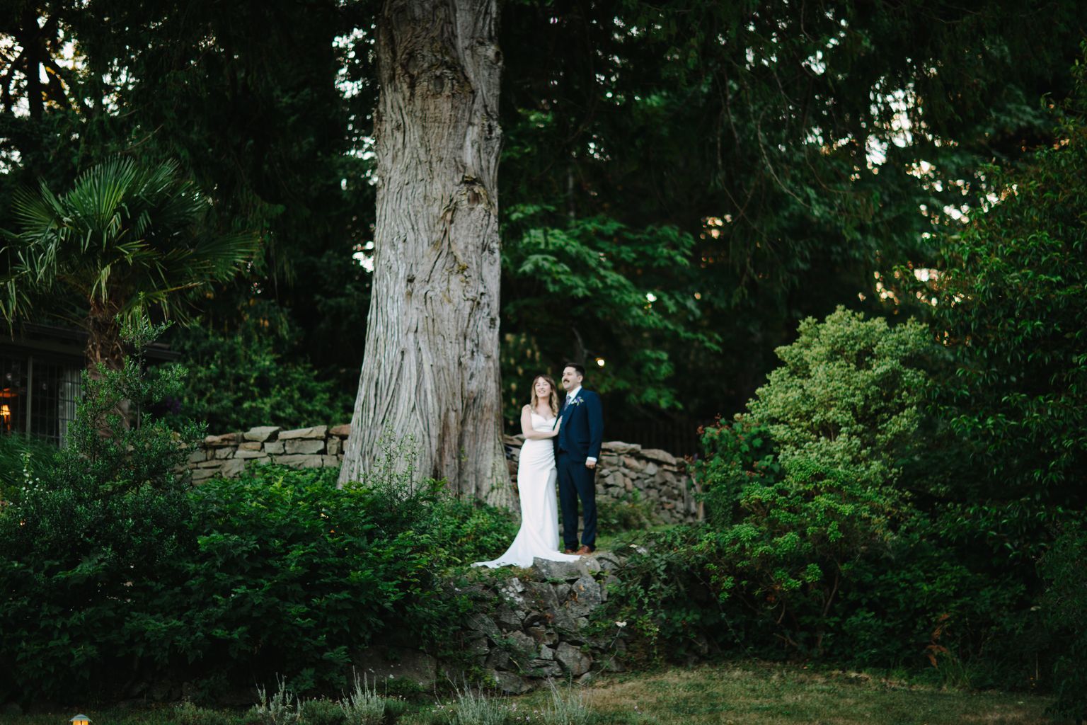 Bride and groom stand together beneath a massive tree trunk at Hastings House, surrounded by lush forest and stone walls