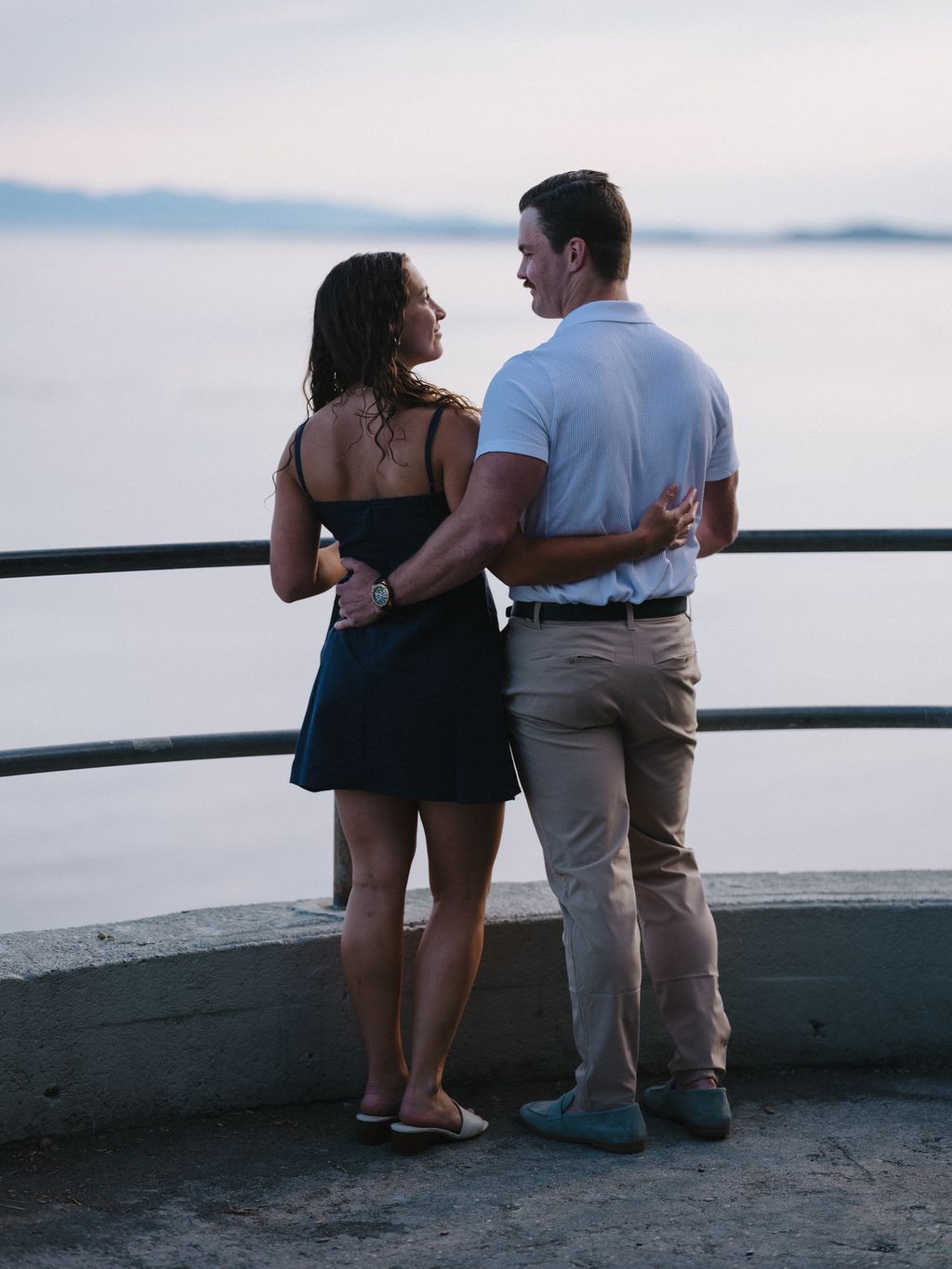 Couple embracing by the water at sunset, woman in black dress and man in blue shirt gazing toward calm coastal waters