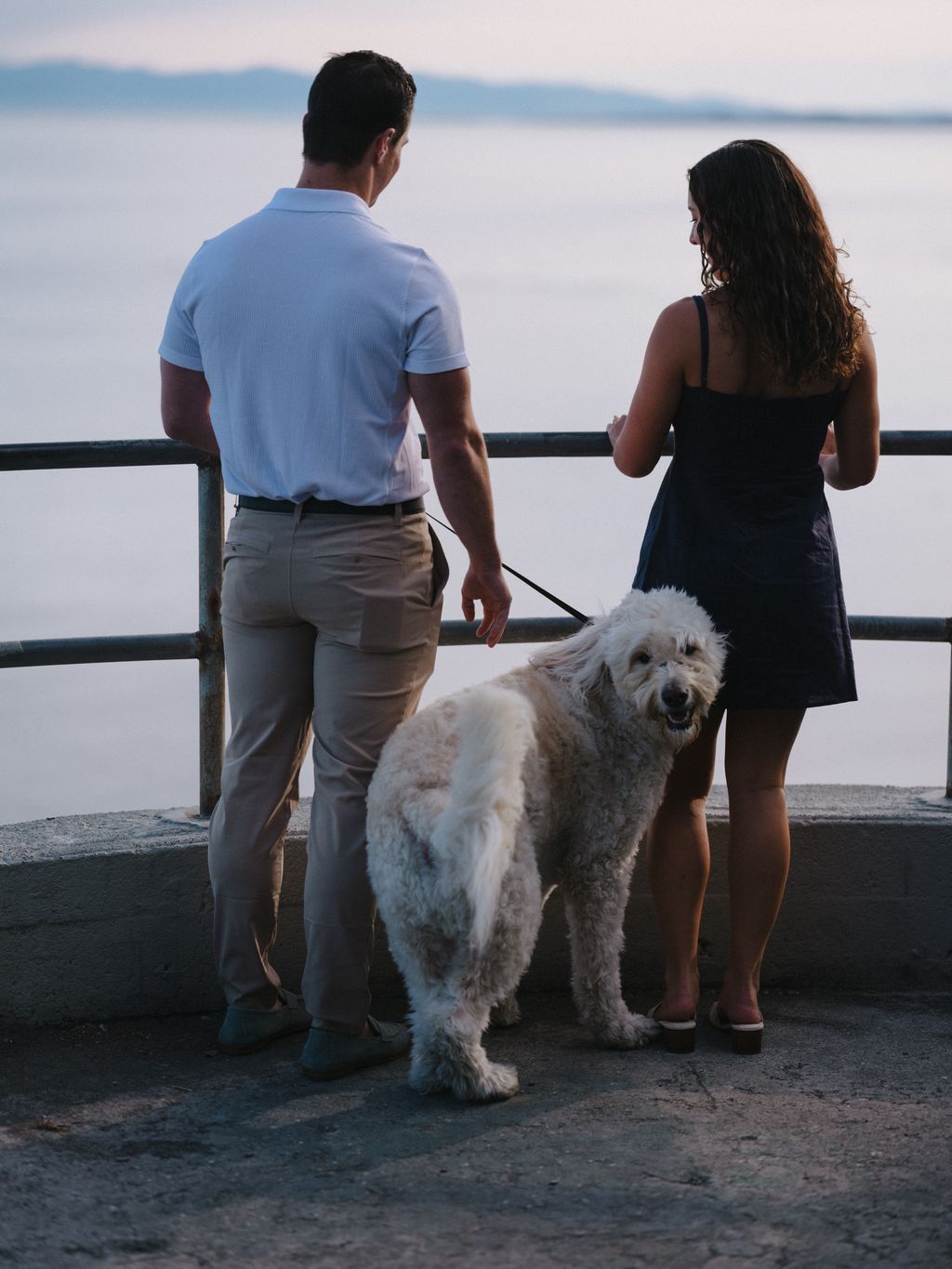 Couple and white dog stand at a wooden railing overlooking calm water at sunset, coastal engagement photography on Salt Spring Island
