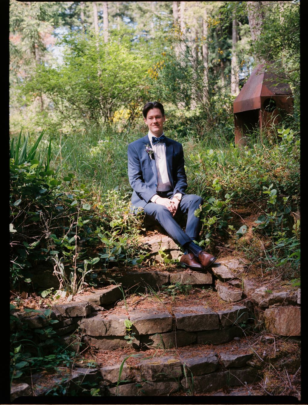 Groom in navy suit and bow tie sits on moss-covered stone steps surrounded by lush forest greenery at Ruckle Park