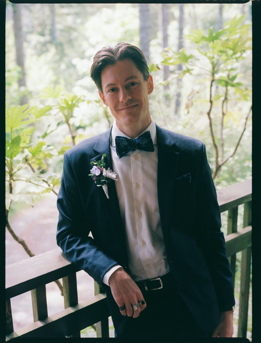 Groom in navy blazer and black bow tie smiles at camera with wildflower boutonnière, evergreen forest soft-focused behind him