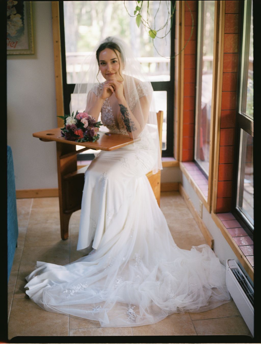 Bride in white lace wedding dress with sheer overlay sits by sunlit window, hand to face, with trailing train and fresh flowers beside her