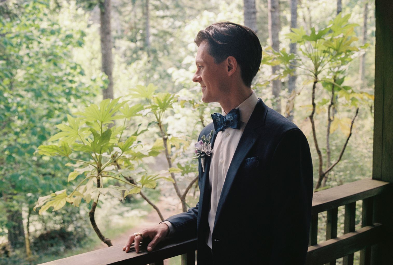 Groom in navy tuxedo and blue bow tie gazes thoughtfully toward blooming white flowers, boutonnière pinned to jacket