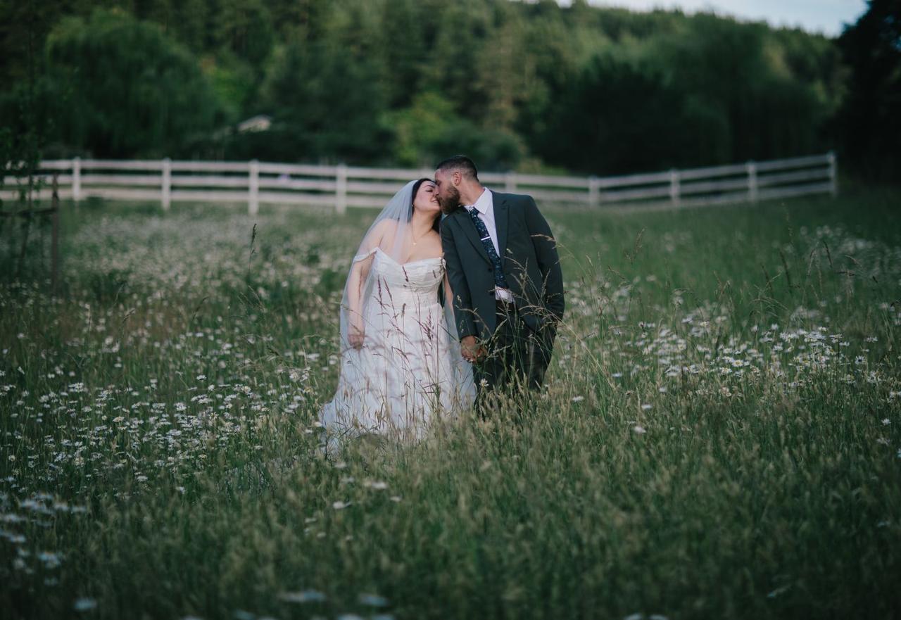 Bride and groom kiss in tall grass field with white fence and evergreen forest backdrop at dusk