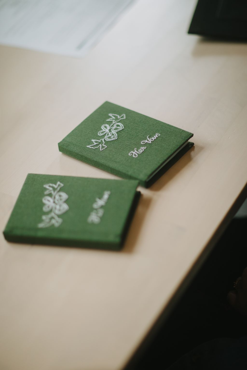Green cloth wedding vows booklets with white embroidered details lie on a neutral surface in soft natural light