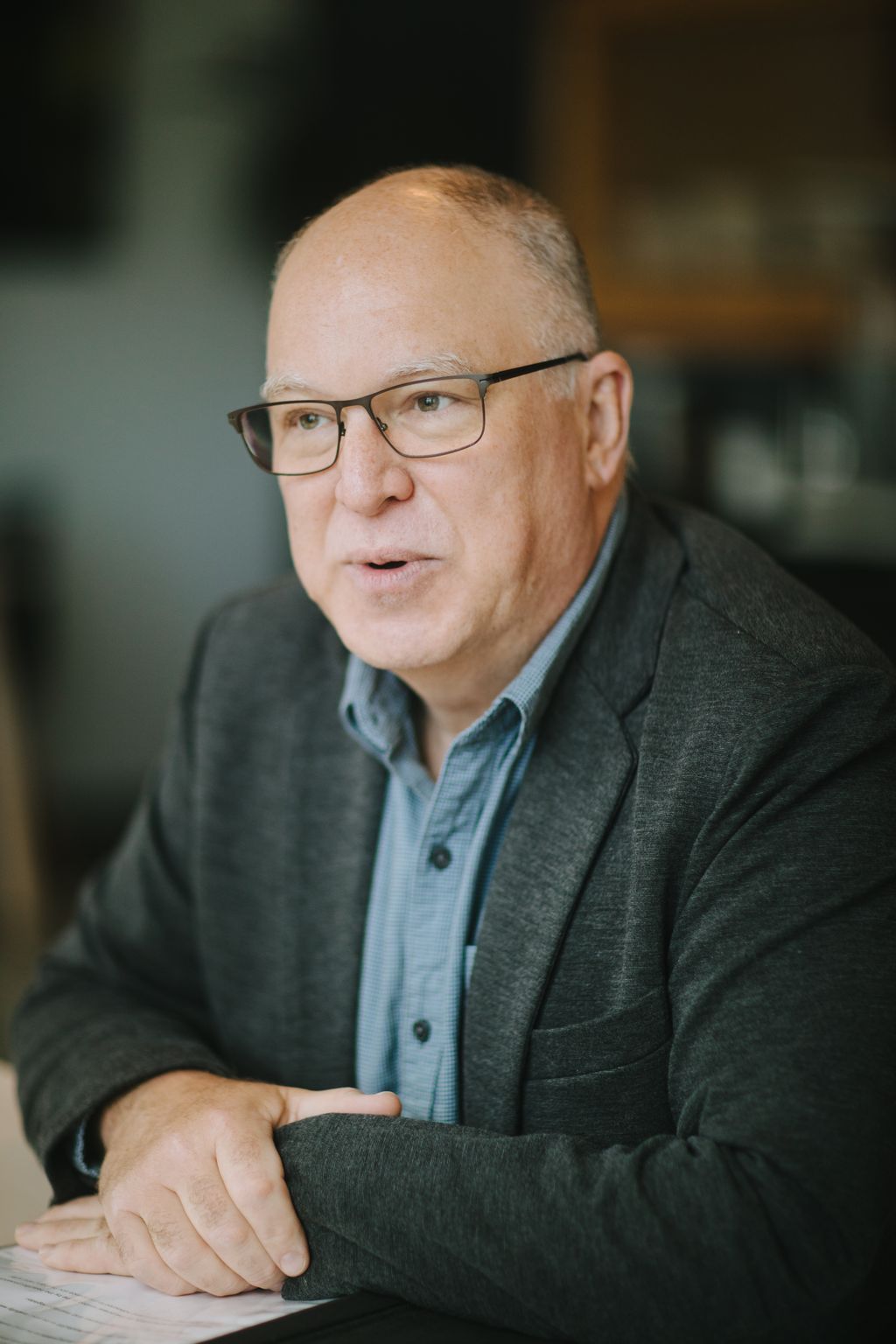 Middle-aged man wearing glasses and gray blazer over light blue shirt, speaking outdoors with blurred natural background