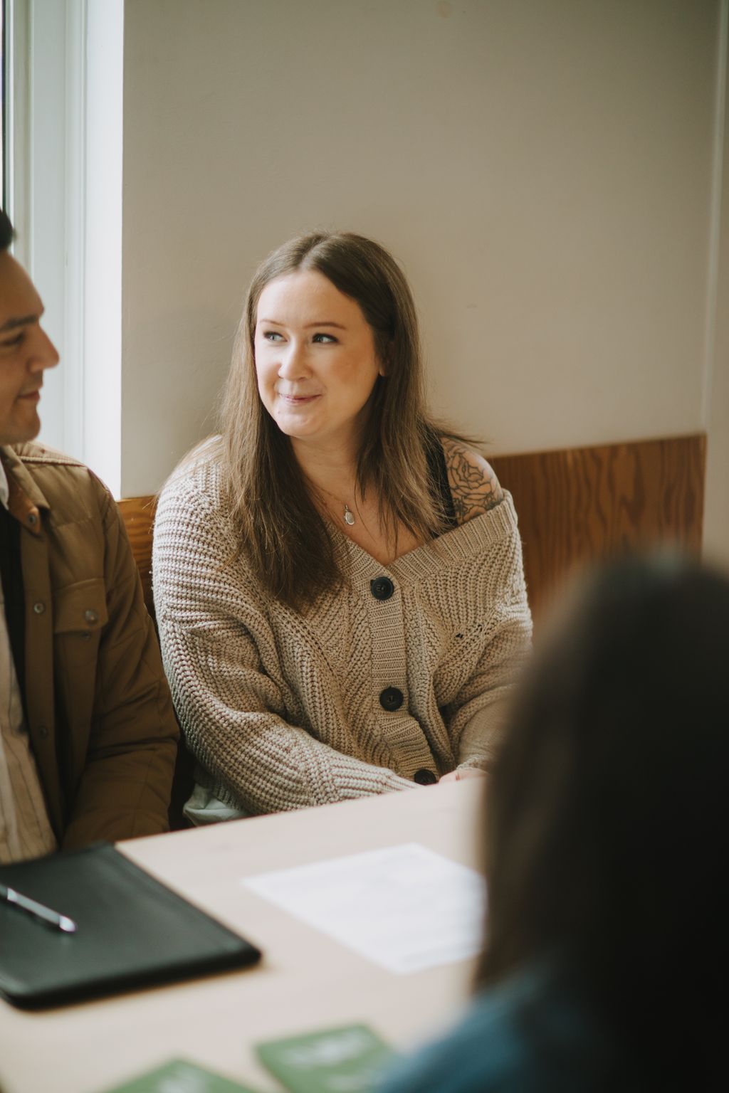 Woman in tan knit cardigan with tattoo on shoulder smiles at table during meeting, warm natural light