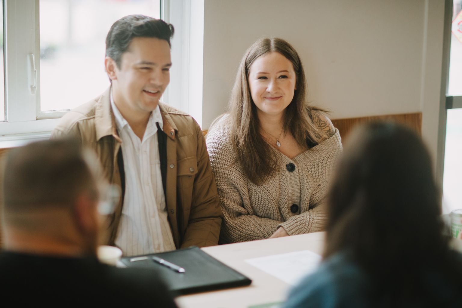 Two people sit at a table, the woman smiling at the camera while the man beside her looks toward her