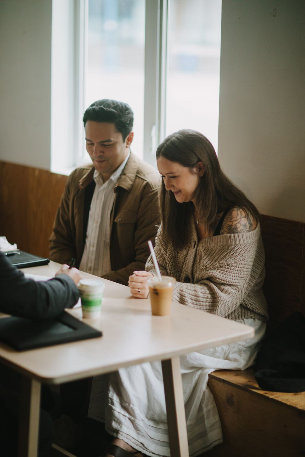 Two people sit at a white table by large windows, holding beverages and looking down with gentle expressions