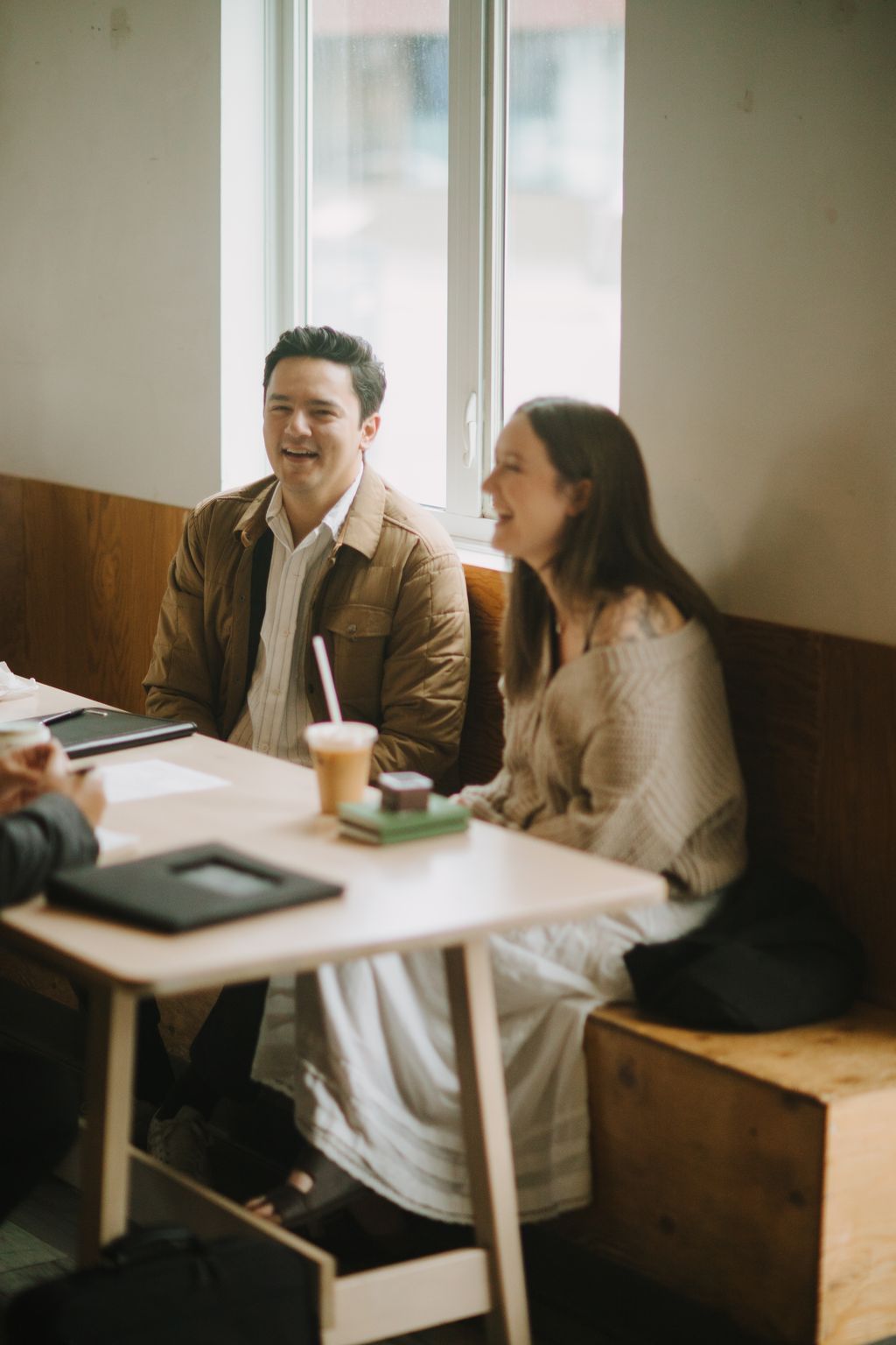 Two people sit at a table by large windows, sharing a laugh over iced drinks in warm afternoon light.