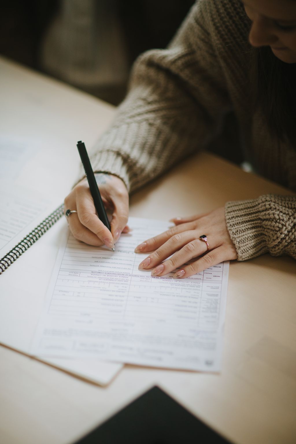 Person wearing a knit sweater and rose gold engagement ring fills out paperwork with a black pen at a wooden table