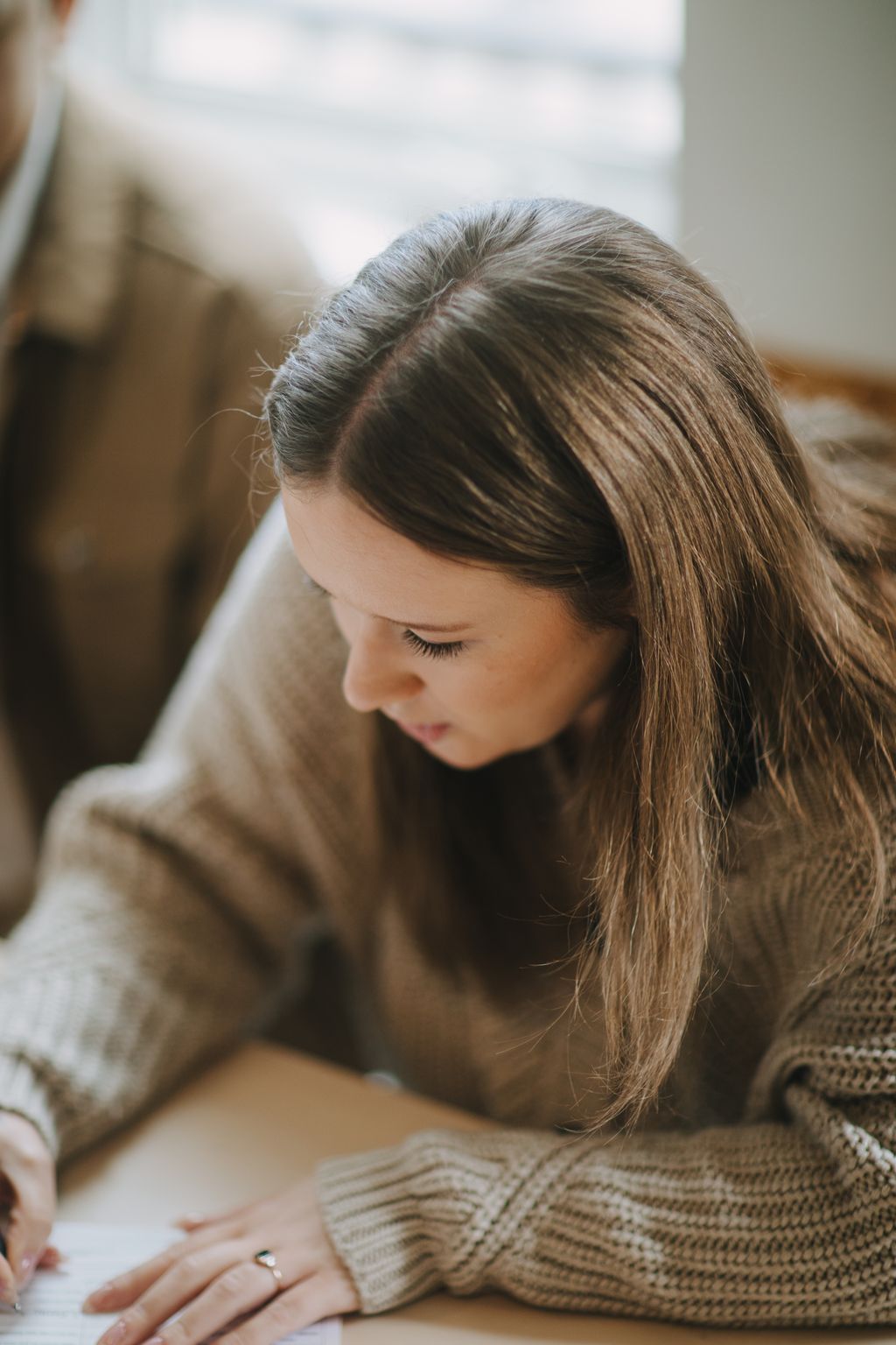 Woman with long brown hair in a side part looking downward, wearing a cream knit sweater, soft natural light outdoors