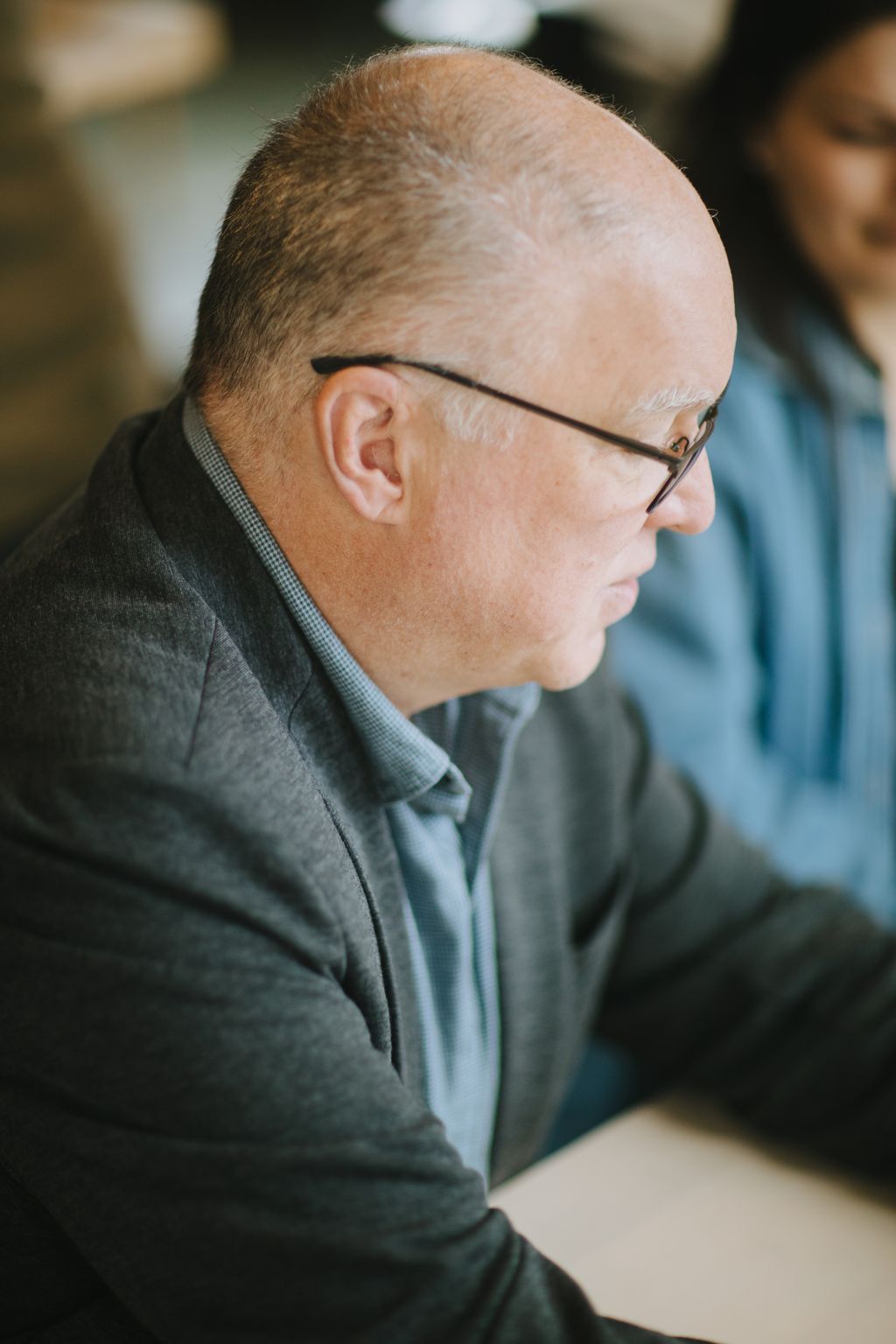 Profile of a bespectacled man in a grey button-up shirt, outdoors in soft natural light