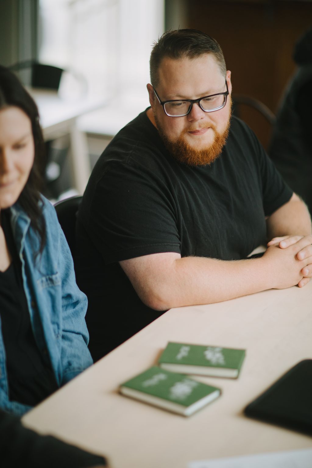 Man with red beard and glasses in black shirt sits at wooden table with green notebooks, looking down thoughtfully
