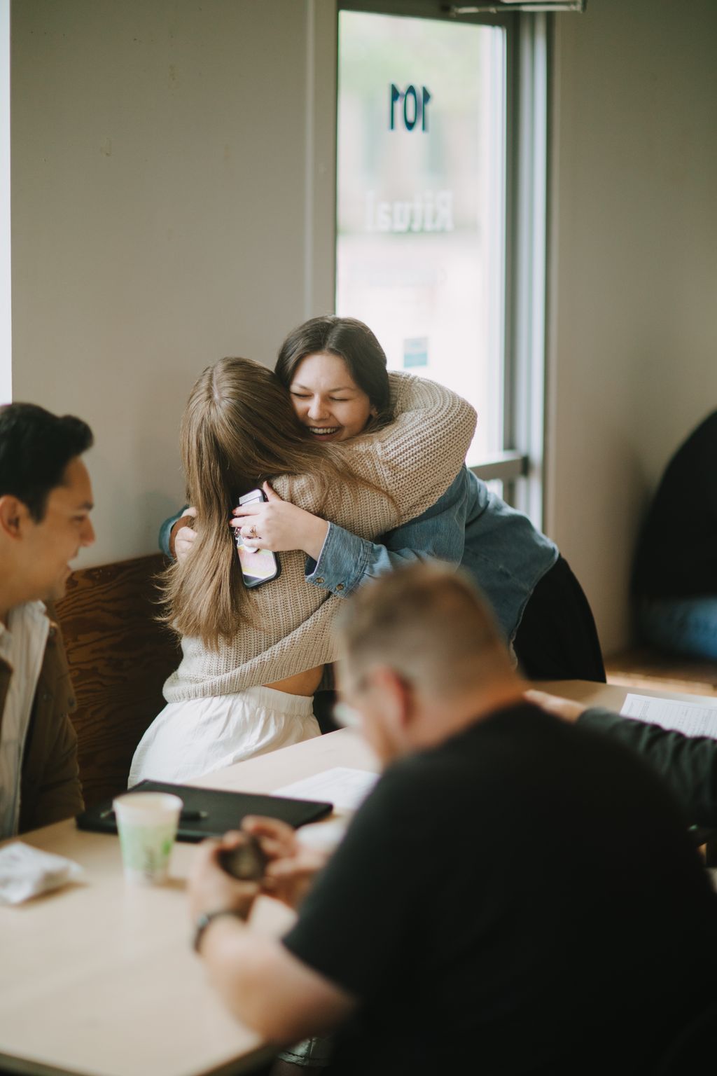 Woman in tan knit sweater hugs person with long hair while smiling, indoor office setting with glass door behind