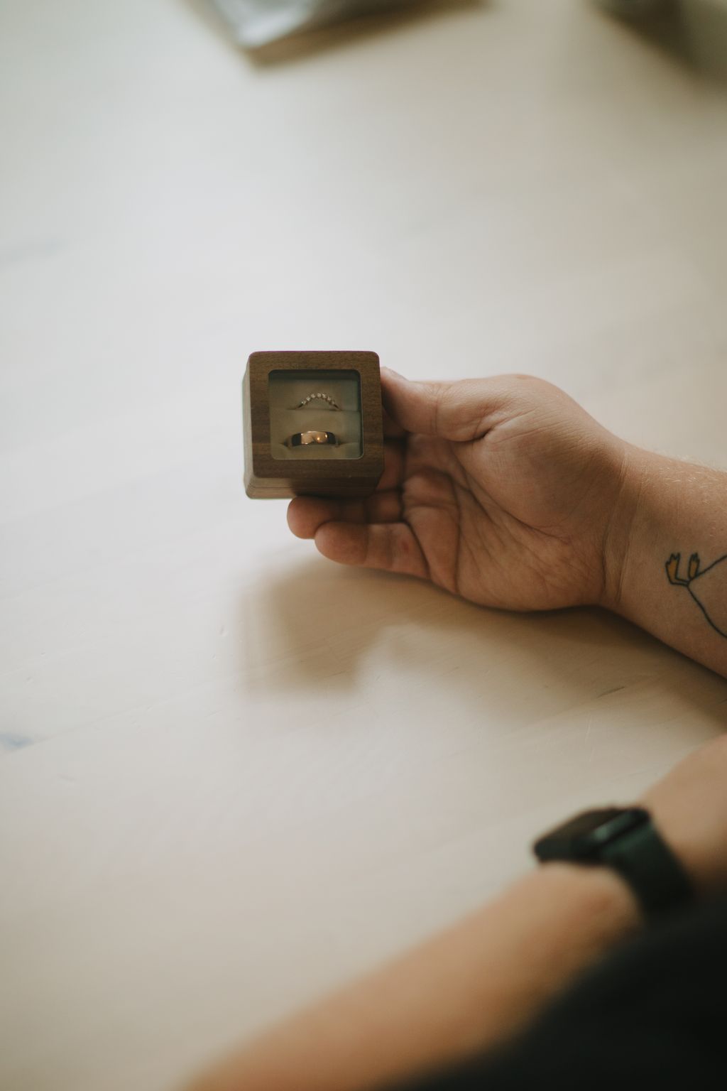 Hand holding open a wooden ring box displaying two gold bands and a diamond arc ring against a neutral background