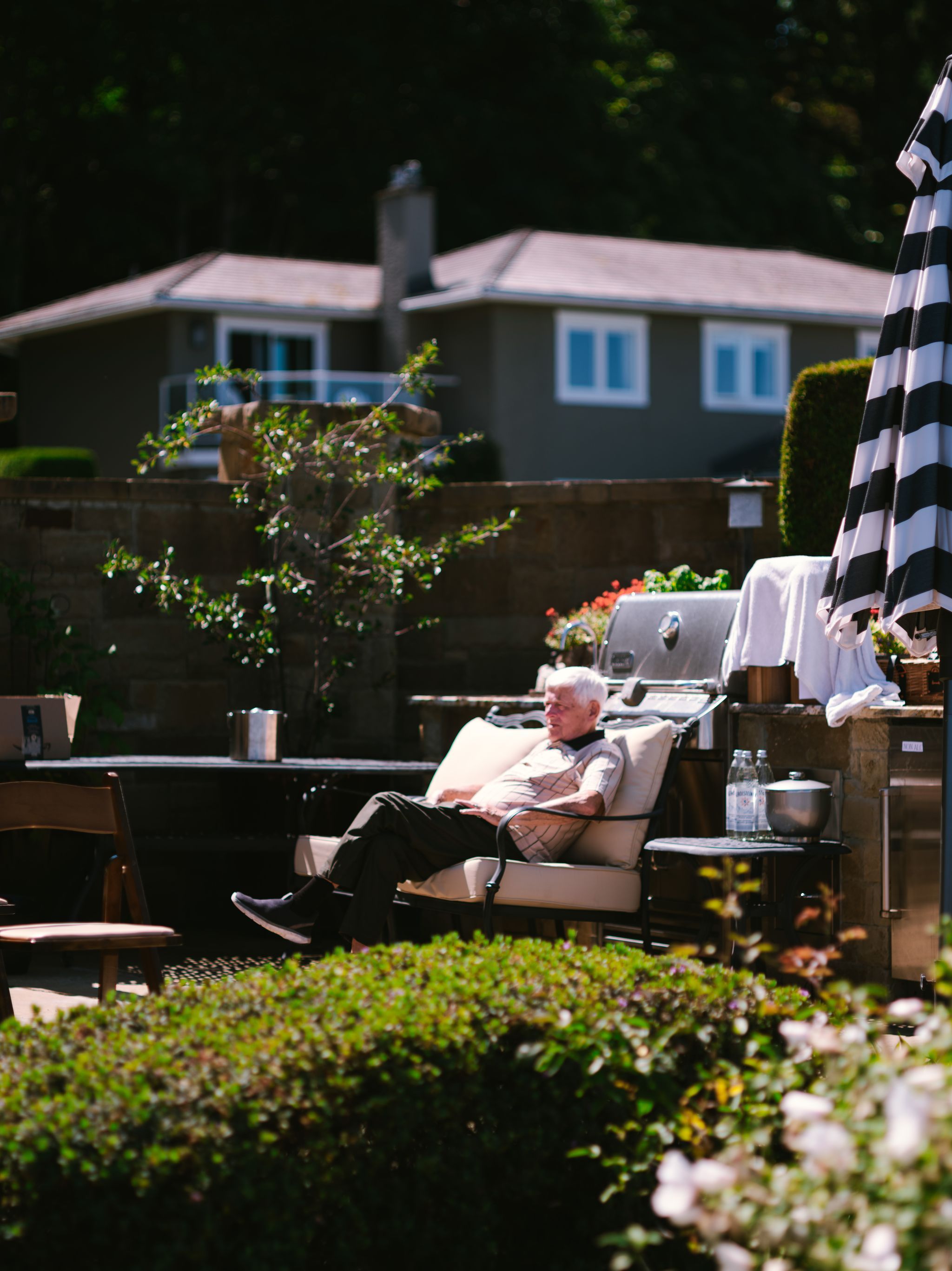 Older man reclines on lounge chair in dappled sunlight, waterfront home and garden visible behind him on Vancouver Island