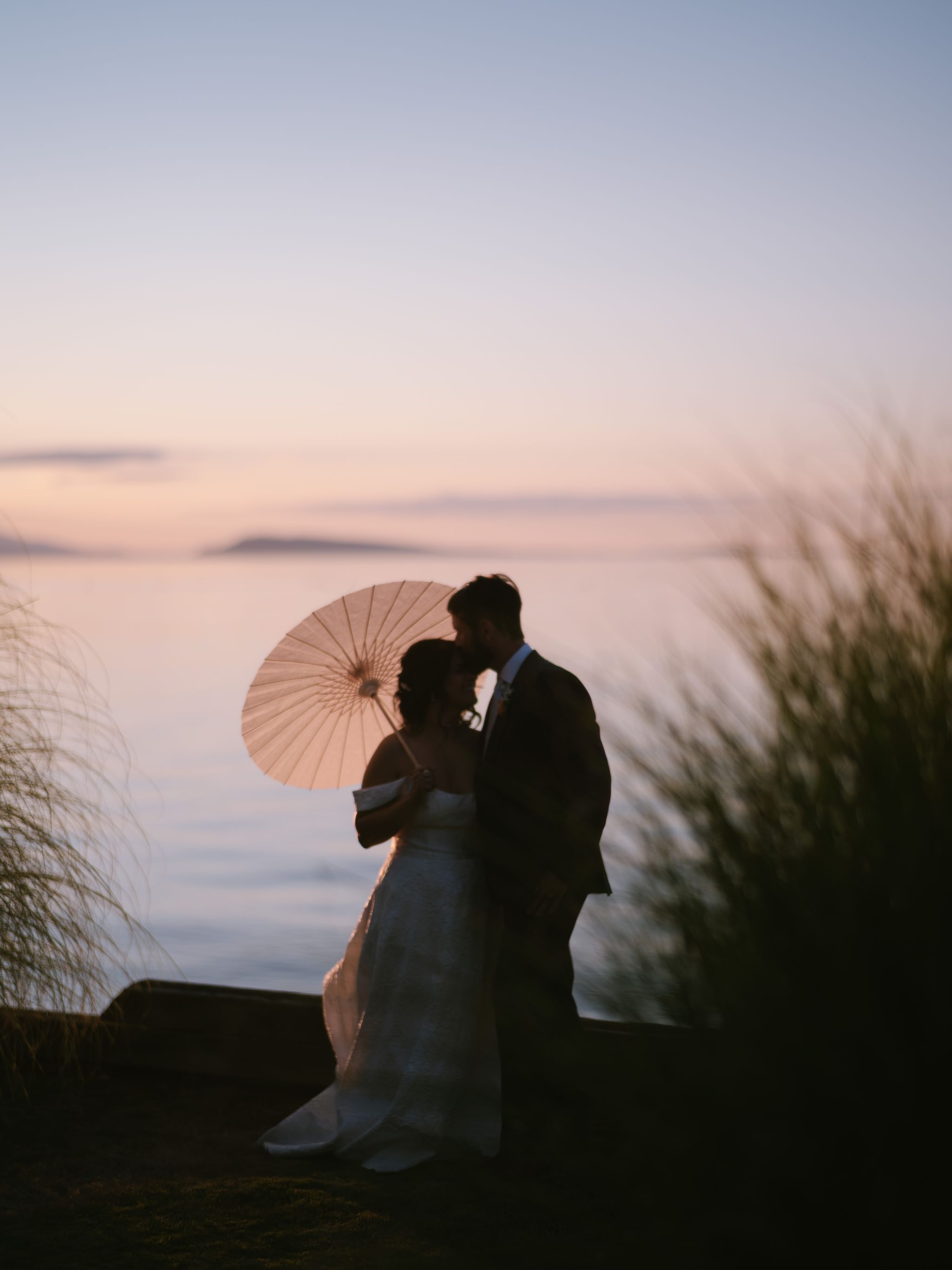 Qualicum Beach wedding couple kissing at sunset, woman holding a parasol, calm water behind