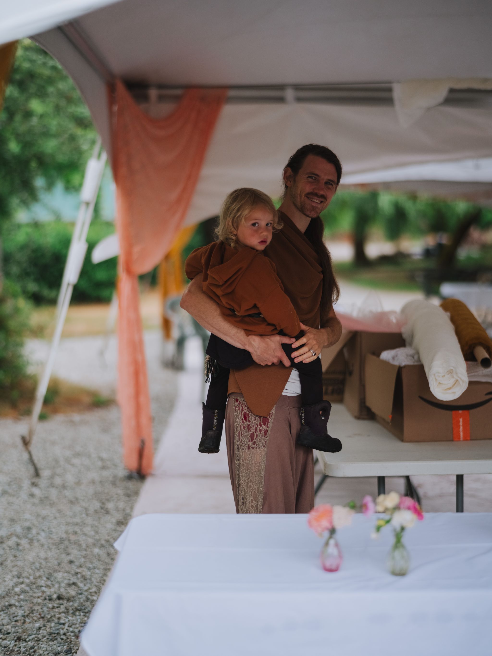 Adult holding toddler in rust-colored wrap at indoor community gathering, soft warm lighting