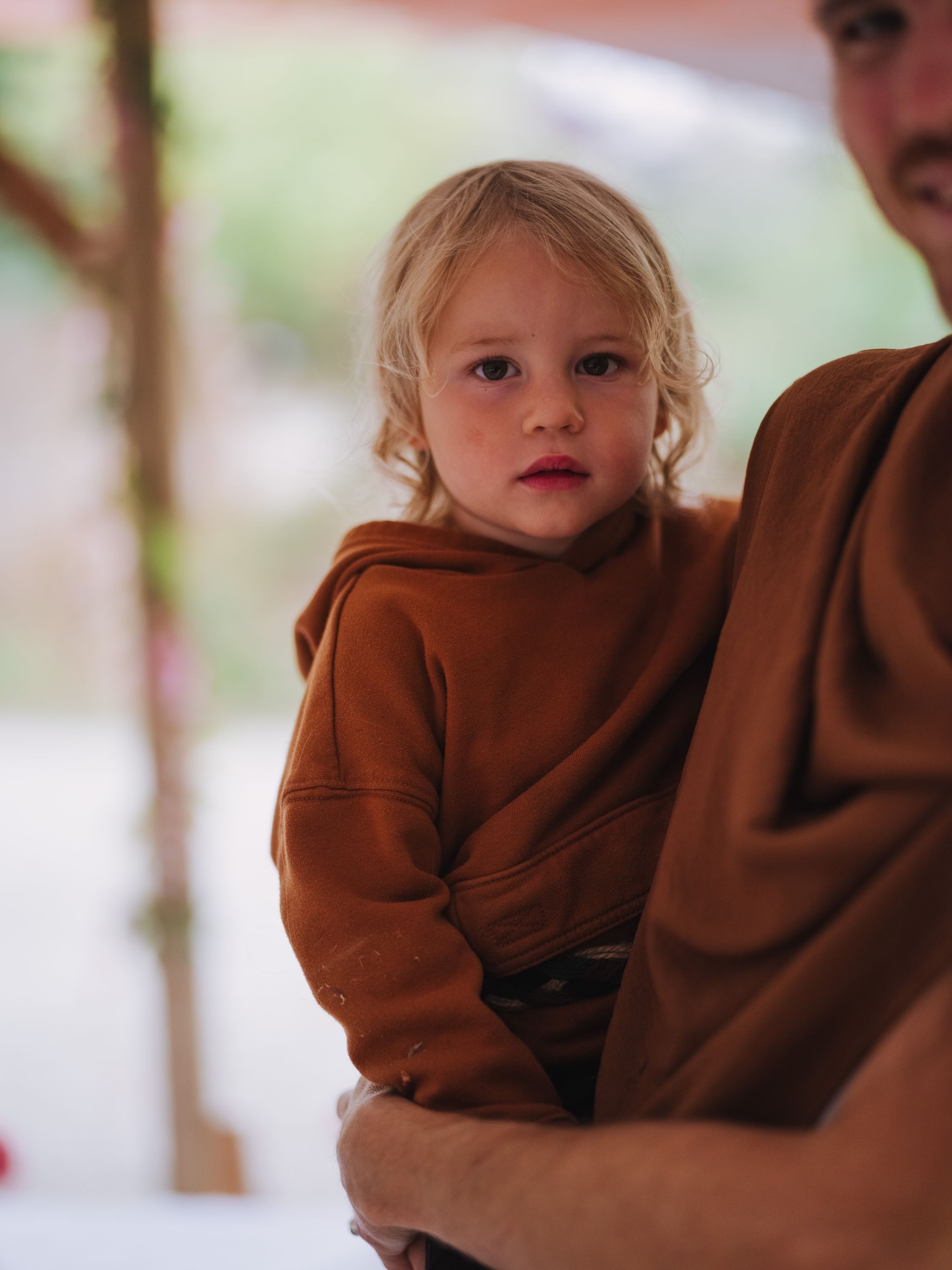 Young child in rust-colored sweater looks over shoulder against soft, blurred pastel background