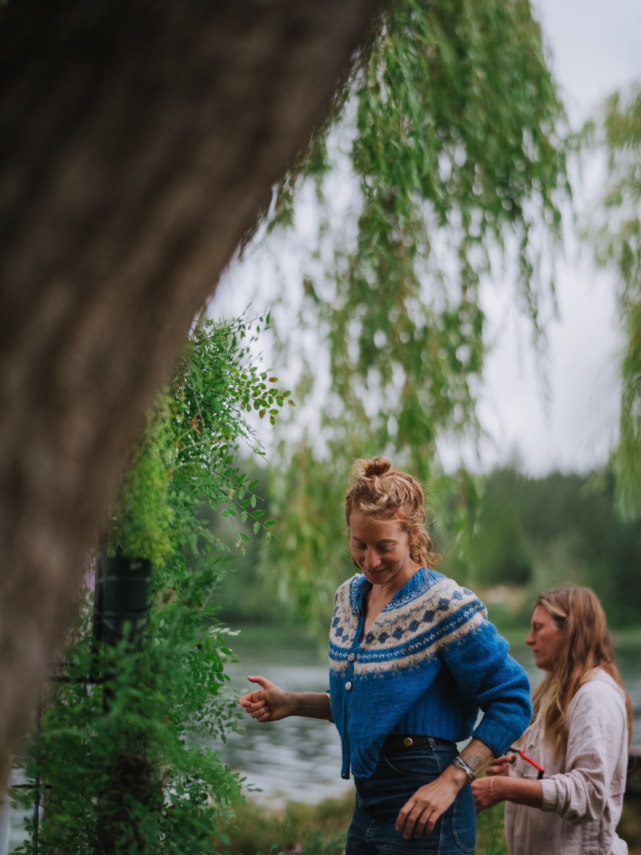 Woman in blue patterned cardigan stands by moss-covered tree beside calm water, hand extended toward hanging branches