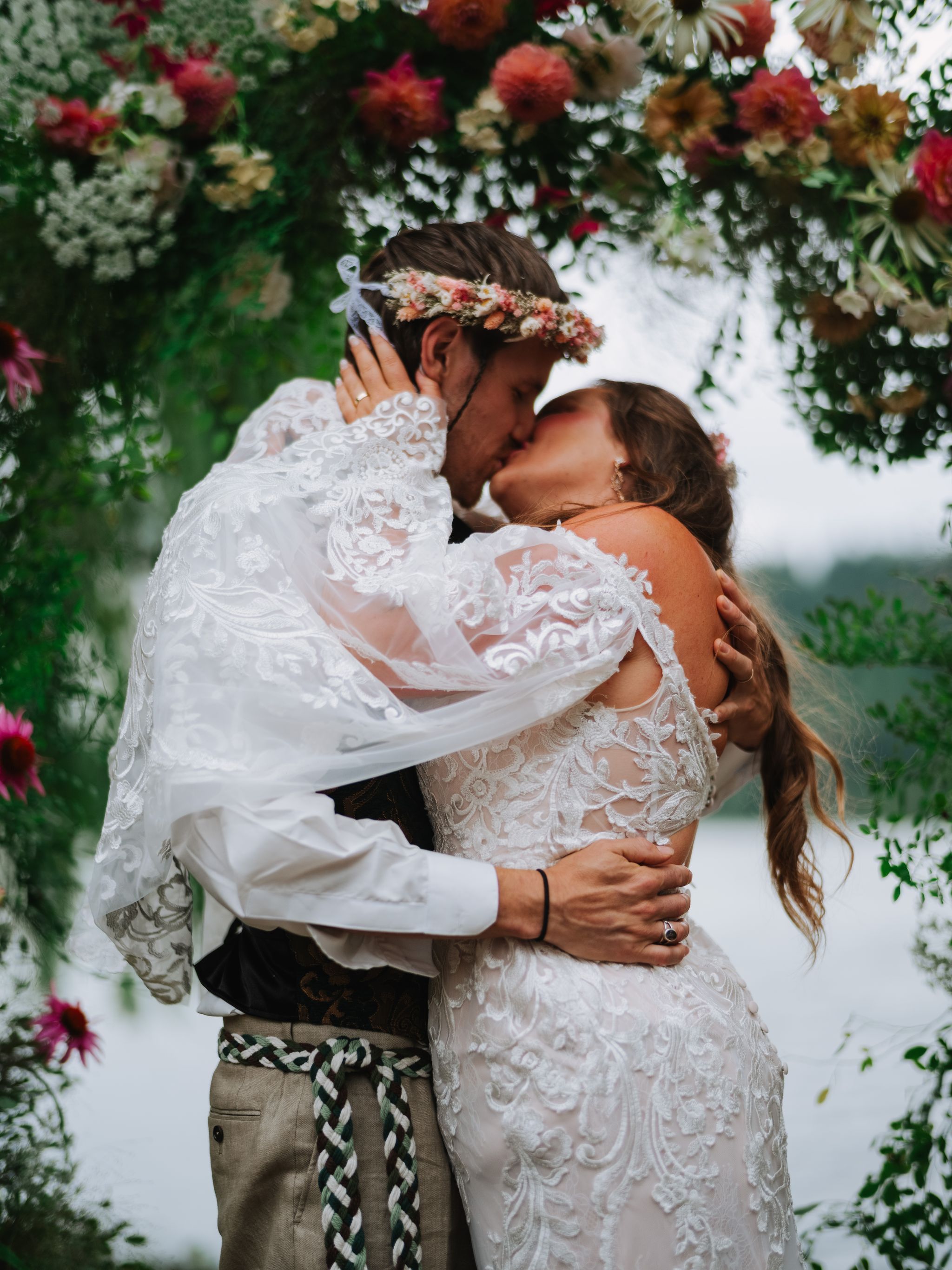 Bride in white lace dress with coral and blush flower crown embraces groom under flowering branches