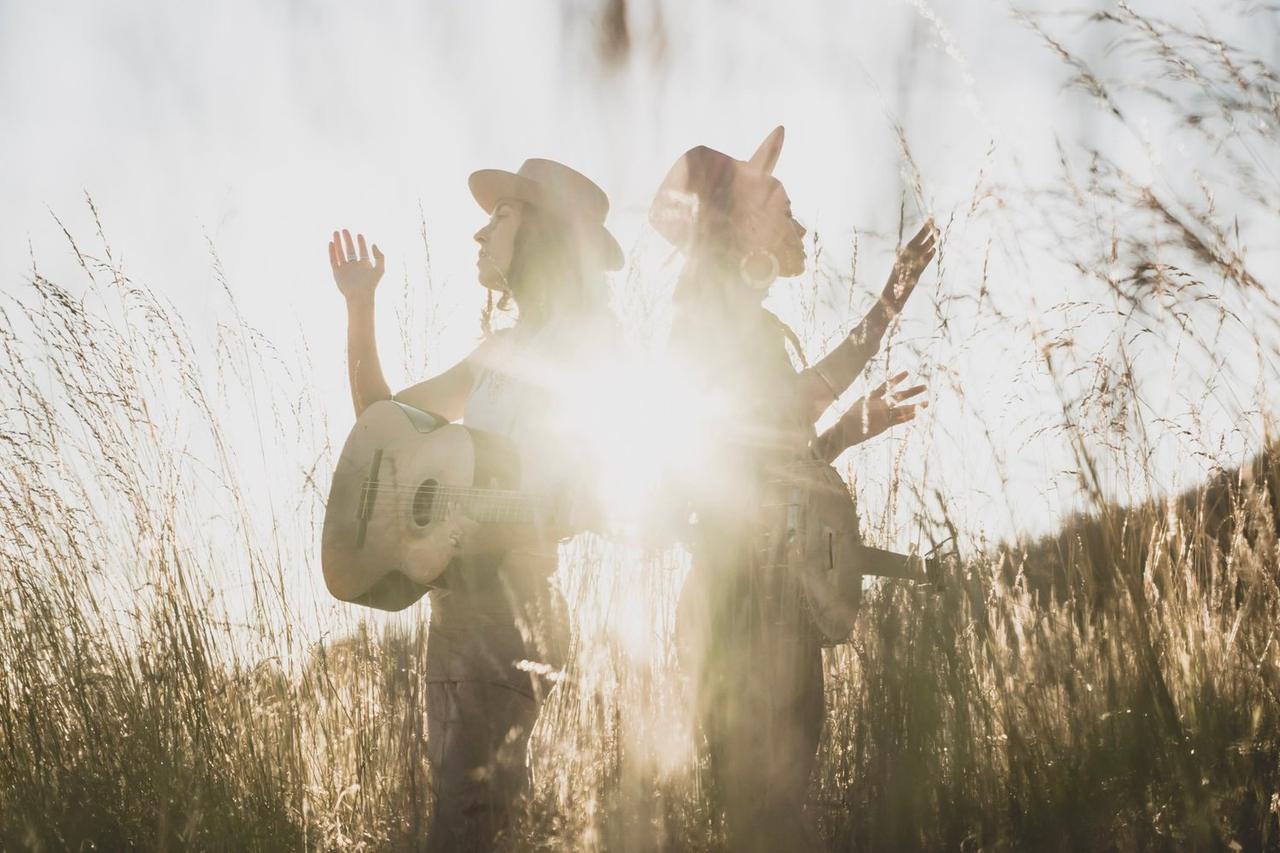 Two figures in cowboy hats stand in dried grass backlit by bright sunlight, faces obscured in silhouette and golden haze